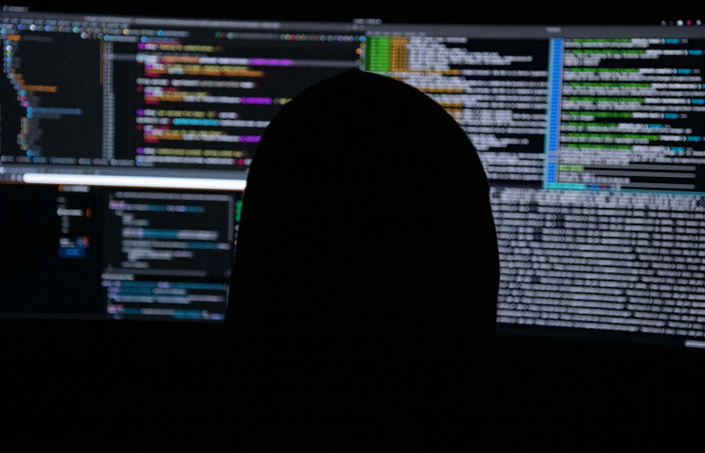 A figure silhouetted against two monitors filled with dense code in a dark room